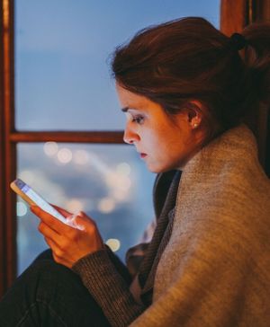zdjęcie przedstawia siedzącą przy oknie kobietę wpatrzoną w ekran smartfona