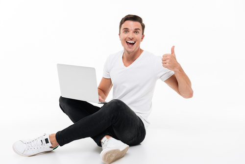 Zadowolony chłopak trzyma na kolanach laptop i pokazuje uniesiony kciuk 