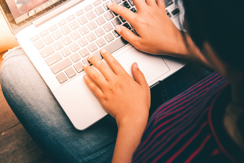 Młoda osoba w ciemnych włosach trzyma na kolanach laptop i pisze na klawiaturze